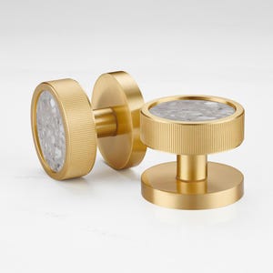 May include: Three gold-toned cabinet knobs with a circular design. Each knob features a textured, ridged edge and a central inset of gray and white speckled stone. The knobs are designed for furniture and home decor.