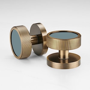 K&ouml;nnte beinhalten: Zwei bronzefarbene Schrankkn&ouml;pfe mit kreisf&ouml;rmigem Design. Jeder Knopf hat einen geriffelten, zylindrischen Au&szlig;enring und eine flache, kreisf&ouml;rmige Basis. Die Mitte jedes Knopfes hat einen blaugr&uuml;nen Einsatz, der einen Kontrast erzeugt.
