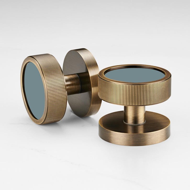 K&ouml;nnte beinhalten: Zwei bronzefarbene Schrankkn&ouml;pfe mit kreisf&ouml;rmigem Design. Jeder Knopf hat einen geriffelten, zylindrischen Au&szlig;enring und eine flache, kreisf&ouml;rmige Basis. Die Mitte jedes Knopfes hat einen blaugr&uuml;nen Einsatz, der einen Kontrast erzeugt.