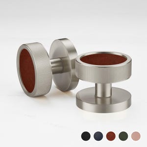 Puede incluir: Un par de gemelos plateados con un borde estriado y una incrustación circular de cuero marrón rojizo. Los gemelos se muestran sobre una superficie blanca, con una selección de muestras de color debajo.