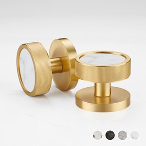 Pode incluir: Dois puxadores de armário dourados com uma inserção de mármore branco. Os puxadores têm um design circular com uma borda canelada. Um puxador é montado numa base redonda, enquanto o outro é um puxador de puxar.