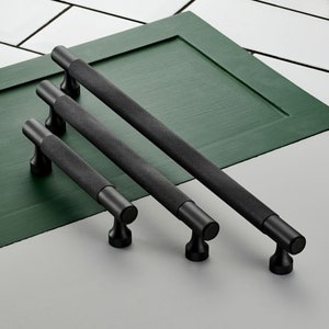 Peut inclure: Trois poignées de tiroir en métal noir avec une finition texturée. Les poignées sont de tailles différentes et sont disposées en rangée sur une surface verte.