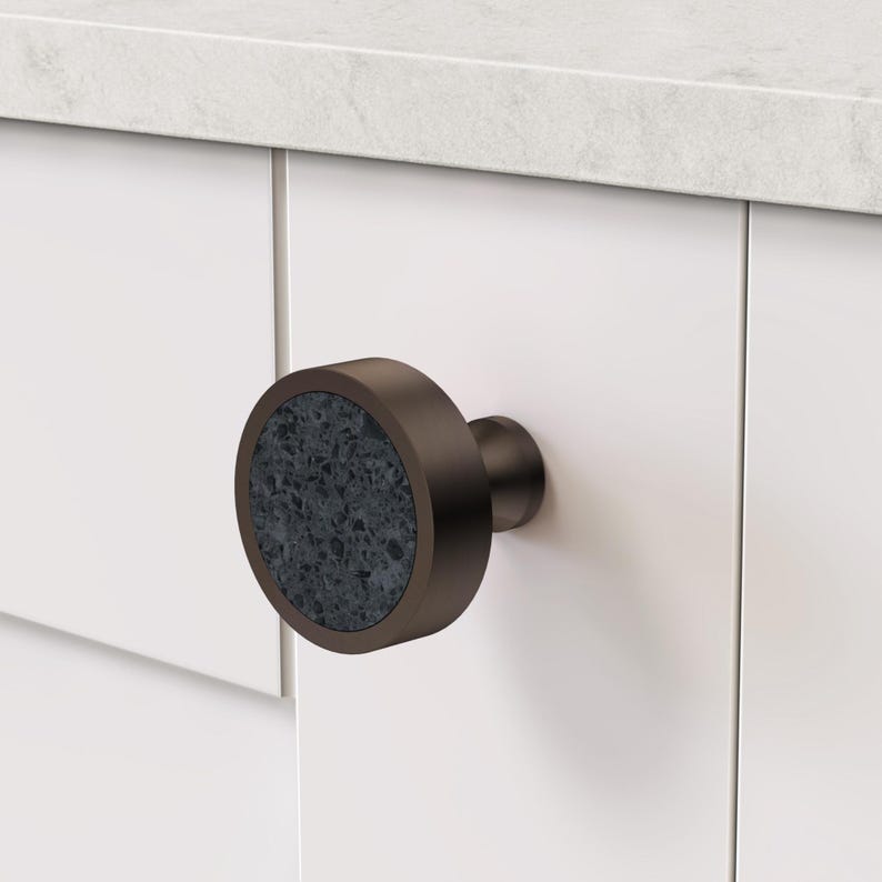 Puede incluir: Un pomo de armario circular con un marco de metal en tono bronce oscuro y una incrustaci&oacute;n de piedra gris oscuro texturizada y moteada. El pomo est&aacute; montado en una puerta de armario blanca, mostrando una base cil&iacute;ndrica y una cara frontal plana.