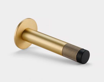 Puede incluir: Un tope de puerta cilíndrico con acabado en oro cepillado, que presenta una placa de montaje circular y un collar metálico texturizado cerca del extremo. La punta del tope está rematada con un protector de goma negro sólido.