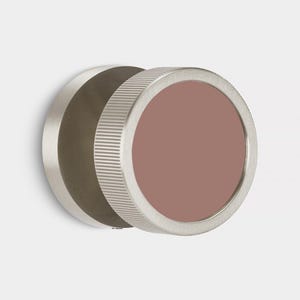 Puede incluir: Primer plano de un objeto circular de metal cepillado con un centro de color malva. El borde exterior tiene una textura estriada y el objeto parece estar montado sobre una superficie blanca. El dise&ntilde;o es minimalista y moderno.