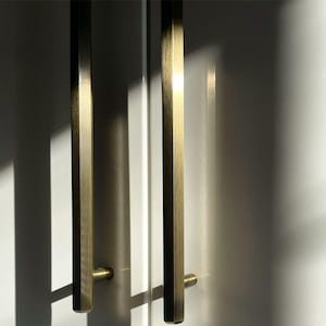 Op de afbeelding: Close-up van twee messingkleurige kastgrepen. De handgrepen zijn rechthoekig met een lichte zeshoekige vorm en zijn verticaal gemonteerd. De achtergrond is een wit oppervlak met schaduwen.