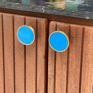 May include: Two circular cabinet knobs featuring a bright blue centre inlay surrounded by a metallic gold-toned rim. The knobs are mounted on vertical-slatted brown wooden cabinet doors with a dark, marbled stone countertop visible above.