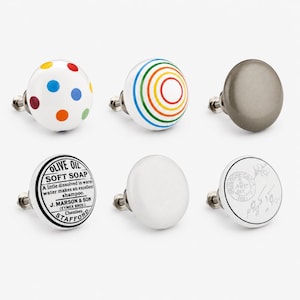 May include: A set of six round cabinet knobs with metal hardware. Designs include a white knob with colorful polka dots, a white knob with rainbow circles, a solid metallic gray knob, a white knob with black text, a solid white knob, and a white knob with a stamp.
