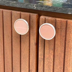 Puede incluir: Dos pomos de armario circulares con un centro rosa claro mate y un borde metálico plateado. Los pomos están montados en puertas de armario de madera con listones verticales que presentan una textura de veta de madera marrón natural.