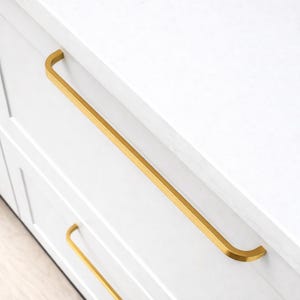 May include: Close-up of white cabinet drawers with gold-colored, rectangular bar pulls. The pulls have a curved design at each end. The cabinet doors are white with a recessed panel design. The countertop is white.