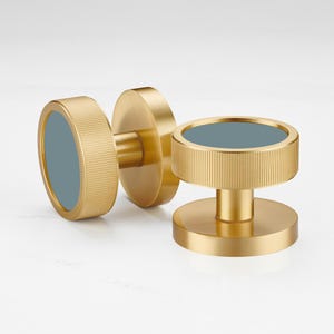 Może przedstawiać: Trzy złote gałki do szafek z żebrowanym wzorem i okrągłym, niebiesko-zielonym środkiem. Gałki są wykonane z metalu i mają nowoczesną, minimalistyczną estetykę. Gałki są przeznaczone do stosowania w szafkach lub szufladach.