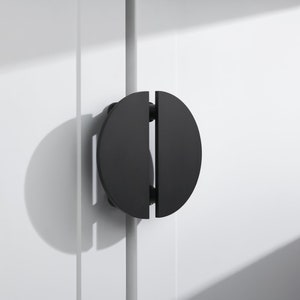 Może przedstawiać: Czarny, okrągły uchwyt do drzwi szafki z podzielonym wzorem. Uchwyt jest wykonany z metalu i ma matowe wykończenie.