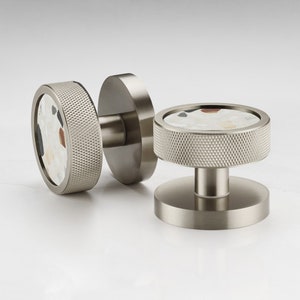 Pode incluir: Dois puxadores de armário em níquel escovado com uma borda texturizada e serrilhada. O centro de cada puxador apresenta um design de terrazzo com manchas brancas, pretas e vermelhas. Um puxador é fixado a uma placa traseira redonda, o outro é independente.