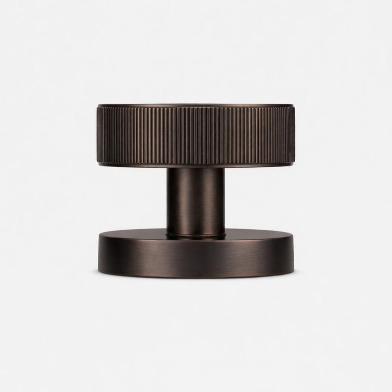 Puede incluir: Un pomo de puerta de color bronce con una parte superior cil&iacute;ndrica estriada y una base circular. El pomo tiene un v&aacute;stago cil&iacute;ndrico corto. Dise&ntilde;o minimalista y moderno.