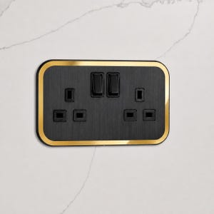 Op de afbeelding: Een rechthoekig dubbel stopcontact met een geborstelde zwarte metalen voorplaat en een gepolijste gouden rand. De unit bevat twee sets driepolige stopcontacten en twee zwarte tuimelschakelaars voor onafhankelijke stroomregeling.
