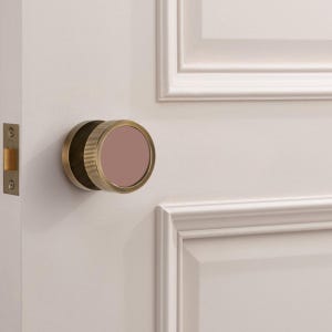 K&ouml;nnte beinhalten: Ein T&uuml;rknauf aus Messing mit einem kreisf&ouml;rmigen, geriffelten Design und einem mauvefarbenen Mittelteil. Der Knauf ist an einer wei&szlig;en T&uuml;r mit passendem T&uuml;rrahmen und einem goldfarbenen T&uuml;rschloss angebracht.