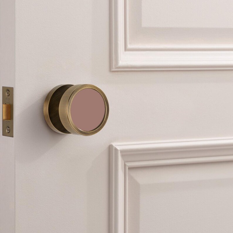 K&ouml;nnte beinhalten: Ein T&uuml;rknauf aus Messing mit einem kreisf&ouml;rmigen, geriffelten Design und einem mauvefarbenen Mittelteil. Der Knauf ist an einer wei&szlig;en T&uuml;r mit passendem T&uuml;rrahmen und einem goldfarbenen T&uuml;rschloss angebracht.