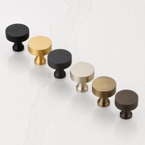 May include: A collection of six circular cabinet knobs arranged in a diagonal line. The knobs feature various metallic and matte finishes, including matte black, brushed gold, polished silver, antique brass, and dark bronze, each with a pedestal base.