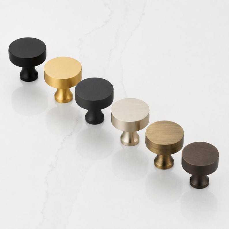 May include: A collection of six circular cabinet knobs arranged in a diagonal line. The knobs feature various metallic and matte finishes, including matte black, brushed gold, polished silver, antique brass, and dark bronze, each with a pedestal base.