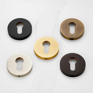 May include: A collection of five round keyhole covers in various metallic finishes: black, brass, gold, brushed nickel, and bronze. Each cover has a keyhole-shaped opening and a small screw hole. The keyhole covers are arranged on a white marble surface.