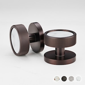 Pode incluir: Dois puxadores de armário de cor bronze com um design circular. Cada puxador apresenta um centro de mármore branco e um anel exterior cilíndrico e estriado. Os puxadores são exibidos sobre um fundo branco.