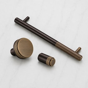 Puede incluir: Un juego de herrajes para armarios que incluye un tirador largo en forma de T, un pomo redondo y un pomo cilíndrico. Cada pieza presenta un diseño bicolor con acabado en bronce oscuro y una sección estriada en tono latón en los extremos.