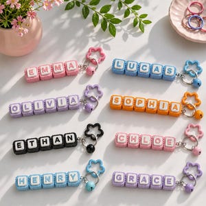 May include: Eight personalized keychains featuring square blocks with white letters spelling the names Emma, Lucas, Olivia, Sophia, Ethan, Chloe, Henry, and Grace. Each keychain includes a metal ring, a small bell, and a flower-shaped charm.