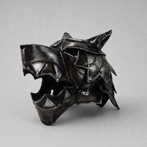 Pode incluir: Um capacete de metal escuro em forma de cabeça de lobo com um design de escamas em camadas, focinho pontiagudo, orelhas afiadas e mandíbula aberta. A superfície tem um acabamento polido e reflexivo com destaques em preto profundo e prata.