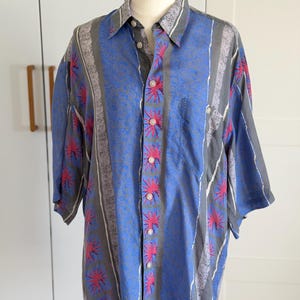 Könnte beinhalten: Ein kurzärmeliges Hemd mit Knopfleiste und vertikalem Streifenmuster in Blau- und Grautönen. Der Stoff zeigt ein wiederkehrendes Design aus leuchtend pinkfarbenen Sternmotiven und dezenten grauen, strukturierten Bändern mit klassischem Kragen.