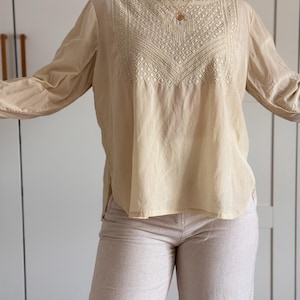 Puede incluir: Una blusa de manga larga color crema con cuello alto y un intrincado bordado tonal en el panel del pecho. La prenda tiene un corte holgado y relajado, mangas largas y un dobladillo recto que incluye pequeñas aberturas laterales en la base.