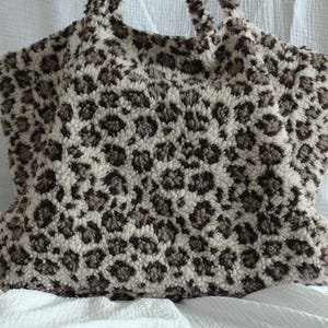Könnte beinhalten: Eine Tragetasche aus einem dicken, flauschigen, cremefarbenen Stoff mit einem dunkelbraunen Leopardenmuster. Die Tasche verfügt über zwei integrierte Griffe und eine weiche, strukturierte Oberfläche, die ein sich wiederholendes Tiermuster zeigt.