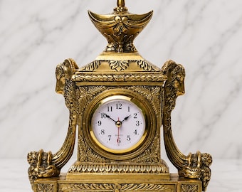 Op de afbeelding: Een goudkleurige schoorsteenklok met een centrale ronde witte wijzerplaat met zwarte Arabische cijfers en een rode secondewijzer. De klok is gevat in een sierlijk gouden frame met gebeeldhouwde ramshoofden en decoratieve bladmotieven.