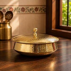 May include: A round, gold-colored metal serving dish with a hammered texture and a matching lid. The lid features a small, decorative knob handle at the center. The dish has a wide, shallow bowl shape with a polished, reflective finish.