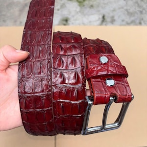 Op de afbeelding: Een donkerrode riem met een gestructureerd, reliëfpatroon dat doet denken aan reptielenschubben. De riem is voorzien van een gepolijste zilverkleurige metalen gesp en twee bijpassende zilverkleurige metalen klinknagels bij de gesp.