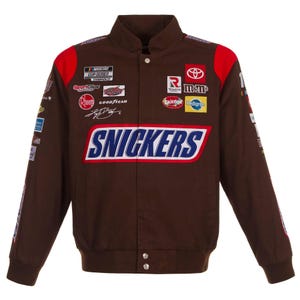 May include: A brown racing jacket featuring a large Snickers logo across the chest. The jacket is adorned with various embroidered sponsor patches including NASCAR Cup Series, Toyota, M&Ms, Skittles, Pedigree, Rheem, and Goodyear, with red shoulder accents.