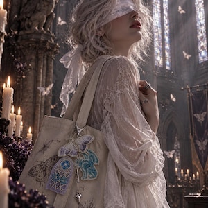 Puede incluir: Un bolso tote de lona beige con un dije de mariposa con cuentas, un parche con motivo de vidriera y un parche azul de forma abstracta. El bolso está adornado con un pequeño dije de corazón plateado y una ilustración de polilla impresa.