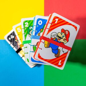 May include: A stack of rectangular playing cards featuring Mario, Luigi, and Yoshi characters, bound together with colorful elastic bands. Each card displays a large number and vibrant graphics, with the top card showing Mario and the number one.
