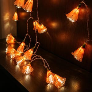 May include: A string of decorative lights featuring translucent orange flower-shaped covers. Each individual blossom houses a small light source that emits a warm glow, connected by a thin, flexible wire that allows for various arrangements.