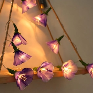 May include: A string of decorative fairy lights featuring translucent purple bell-shaped flower blossoms. Each flower contains a small warm white LED bulb at its center, connected by a thin wire, creating a soft glowing illumination effect.