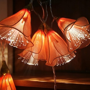 May include: A string of decorative lights featuring multiple orange, bell-shaped flower covers. Each flower is constructed from a translucent, textured fabric and contains a cluster of thin, illuminated fiber optic strands that emit a bright white glow.