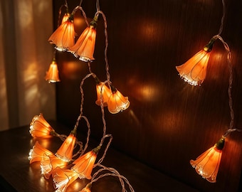 May include: A string of decorative lights featuring translucent orange flower-shaped covers. Each individual blossom houses a small light source that emits a warm glow, connected by a thin, flexible wire that allows for various arrangements.