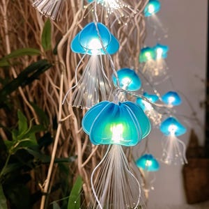 May include: A string of decorative LED lights shaped like jellyfish, featuring translucent blue bell-shaped caps and long, thin, white fibre optic tentacles that glow from within, connected by a thin, flexible, transparent electrical wire.