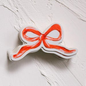 Può includere: Una pila di adesivi con un design a fiocco rosso con contorni bianchi. L'arco ha un effetto acquerello, con ombre e luci. Gli adesivi sono su una superficie bianca strutturata.