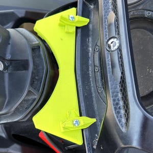 Puede incluir: Un soporte de plástico amarillo neón brillante en forma de V, montado entre una consola de moto acuática negra y un conjunto de manillar negro. El soporte está asegurado con dos tuercas de mariposa y pernos metálicos plateados.