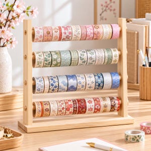 Könnte beinhalten: Ein helles Holzregal mit vier horizontalen Stangen, an denen mehrere Rollen dekoratives Washi-Tape hängen. Die Bänder sind nach Farben in Reihen angeordnet und zeigen florale sowie geometrische Muster in Rosa, Grün, Blau und Rot.