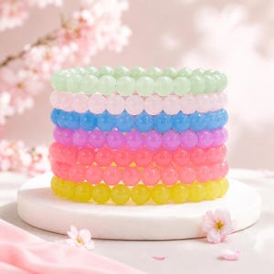 May include: A stack of seven elastic bracelets made of smooth, round, polished beads in a rainbow of colours including mint green, pale pink, sky blue, lavender, coral, bright pink, and yellow, arranged in a vertical column on a white base.