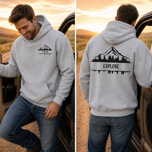 Könnte beinhalten: Ein hellgrauer Kapuzenpullover mit einem schwarzen Grafikdruck, der ein Gebirge, Tannenbäume und das Wort EXPLORE zeigt. Die Vorderseite hat ein kleines Brustlogo, während die Rückseite eine größere Version desselben Bergmotivs aufweist.