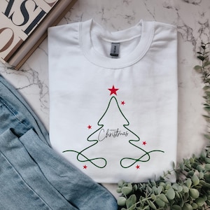 Könnte beinhalten: Ein weißes T-Shirt mit Rundhalsausschnitt und einer minimalistischen, grünen Linienzeichnung eines Weihnachtsbaums. Das Design enthält kleine rote Sterne und den schwarzen, kursiven Schriftzug "Merry Christmas" in der Mitte des Baumes.