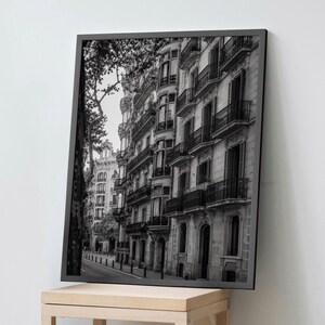 May include: A black and white framed wall art print depicting a street-level view of a multi-storey European building with ornate balconies, tall windows, and architectural detailing, positioned on a light-coloured wooden display stand.
