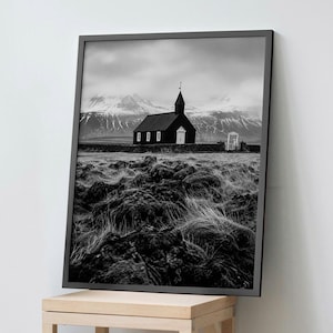 May include: A black and white framed wall art print featuring a dark church with a steeple, situated in a field of rocky terrain and tall grass, with snow-capped mountains under a cloudy sky in the background, presented in a thin black frame.